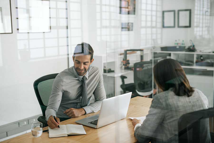 AdobeStock_267740448 Man and woman in office writing and talking with laptop on desk