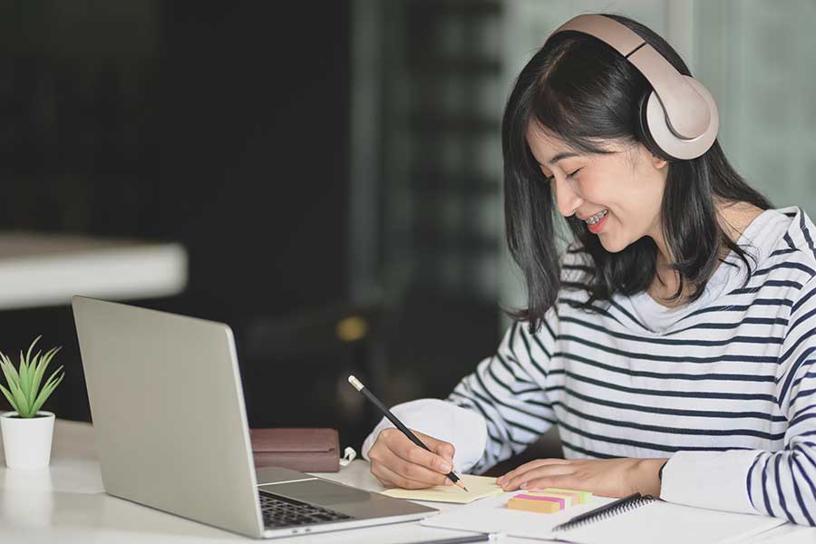 Asian woman writing on notepad with laptop and headphones.