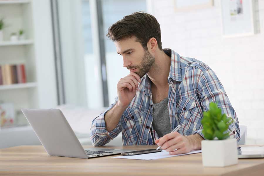AdobeStock_77080791 Young man writing copy on notapad while looking at laptop.