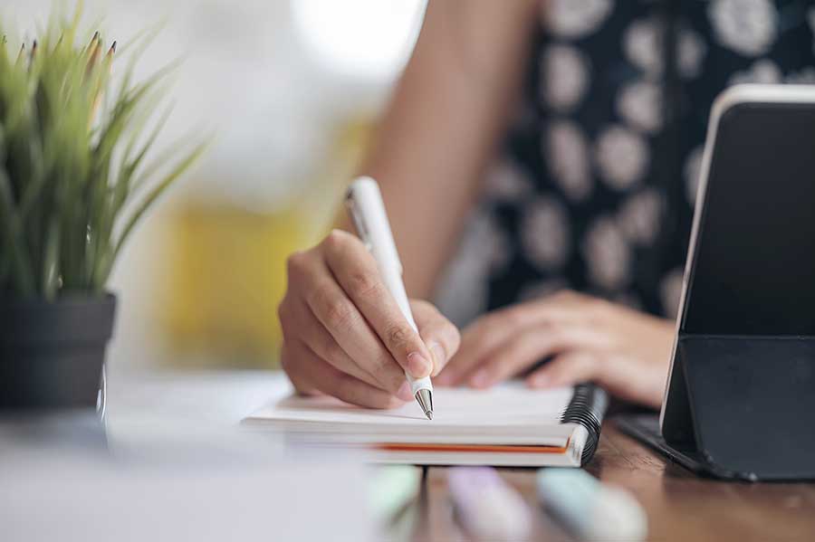AdobeStock_332387392 Woman's hand writing on a notepad beside laptop