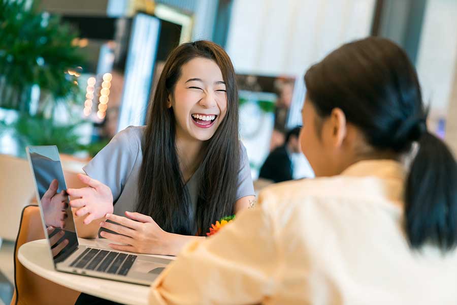 Two Japanese women talking at a desk looking at a laptop.