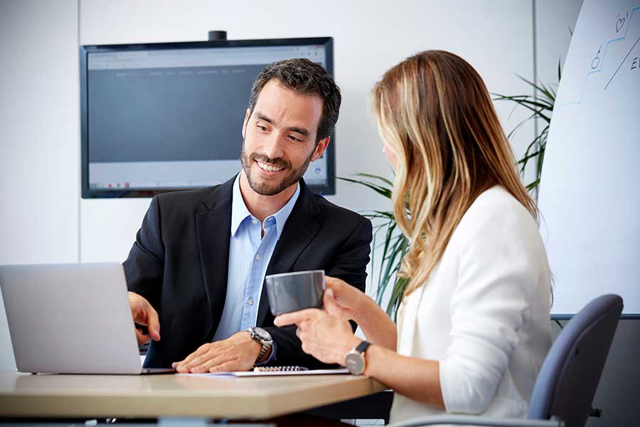 Man and women in an office sitting at a deck looking at a computer. Man is smiling and woman has a cup of coffee.