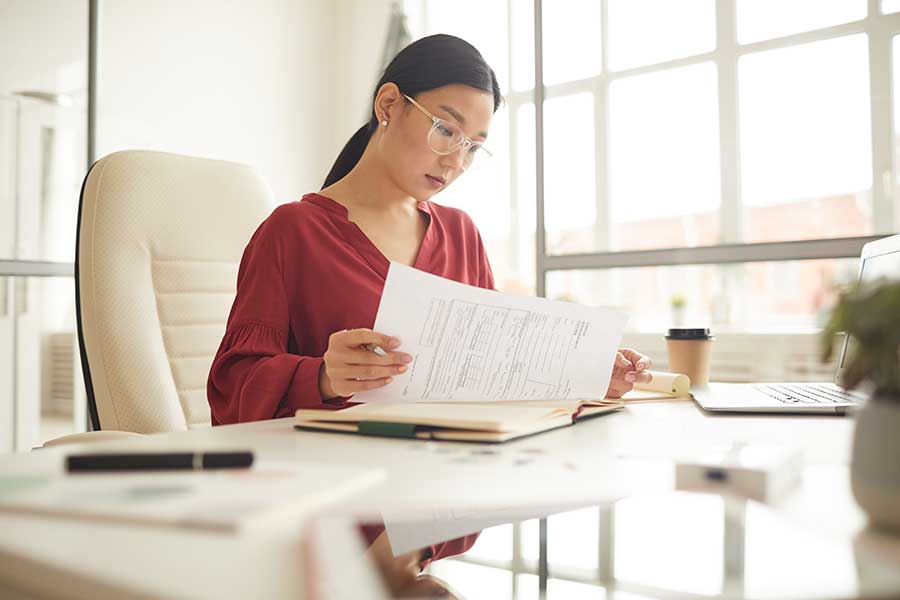 Femme asiatique avec des lunettes lisant des documents à un bureau dans un bureau brillamment éclairé. 