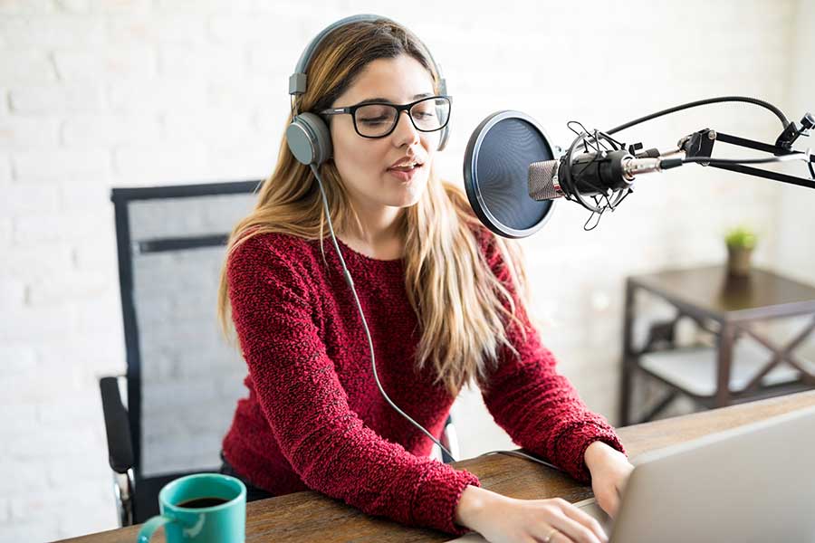 Woman recording into a microphone with headphones on. She is at a desk wearing a jumper and has glasses on. 
